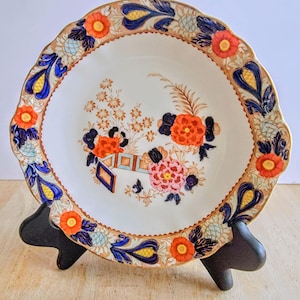 May include: A decorative ceramic plate with a scalloped edge, featuring a floral design in orange, blue, and pink. The plate has a gold trim and is displayed on a black stand. The plate's design includes flowers and geometric shapes.