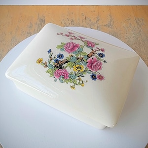 May include: A white ceramic box with a floral design. The design features pink, yellow, blue, and green flowers on a branch. The box has a lid and is rectangular in shape.