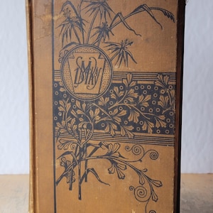 May include: A vintage hardback book with a tan cloth cover featuring black ink illustrations of ferns and foliage. A circular emblem on the front contains the text "Lorna Doone" in a stylised script. The book has a rectangular shape and worn edges.