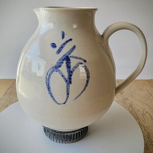 May include: Cream-colored ceramic pitcher with a rounded body and a curved handle. The pitcher features a blue abstract design on the front. It sits on a dark metal base. The pitcher is suitable for serving beverages.