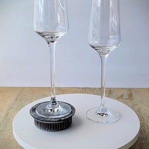 May include: Two clear champagne flutes with long stems. One flute rests on a dark gray floral frog, the other on a white circular surface. The flutes are empty and have a simple, elegant design.
