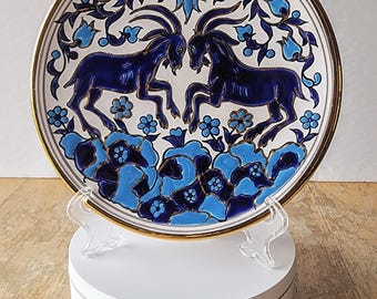 May include: A white ceramic plate with a blue and gold design of two goats facing each other. The plate is decorated with floral motifs and has a gold rim.