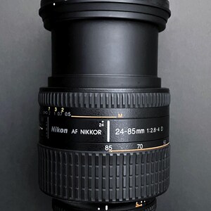 May include: Black Nikon AF Nikkor 24-85mm 1:2.8-4 D camera lens. The lens has a ribbed focus ring and aperture settings from 2.8 to 22. The lens has a focal length range of 24-85mm.