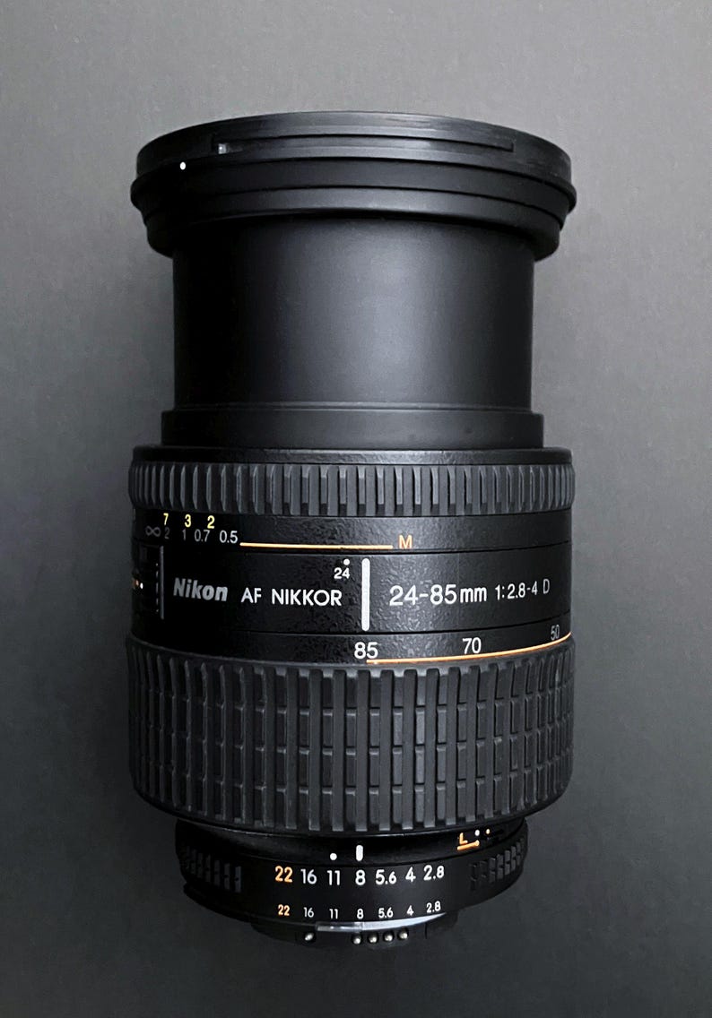 May include: Black Nikon AF Nikkor 24-85mm 1:2.8-4 D camera lens. The lens has a ribbed focus ring and aperture settings from 2.8 to 22. The lens has a focal length range of 24-85mm.