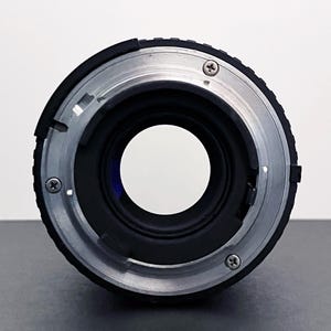May include: Close-up of a camera lens mount. The lens has a black body with a silver-colored metal ring. Three small screws are visible on the silver ring. The lens opening is a large, clear circle.