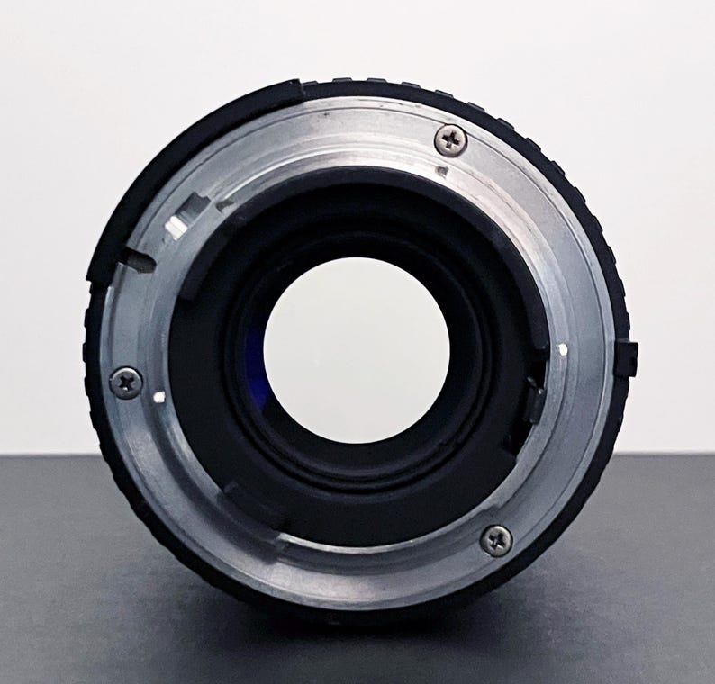 May include: Close-up of a camera lens mount. The lens has a black body with a silver-colored metal ring. Three small screws are visible on the silver ring. The lens opening is a large, clear circle.