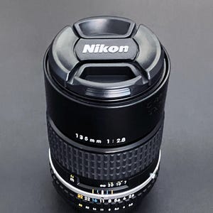 May include: Black Nikon 135mm f/2.8 camera lens with a lens cap. The lens has a ribbed focus ring and aperture settings. The lens cap has the Nikon logo in white.