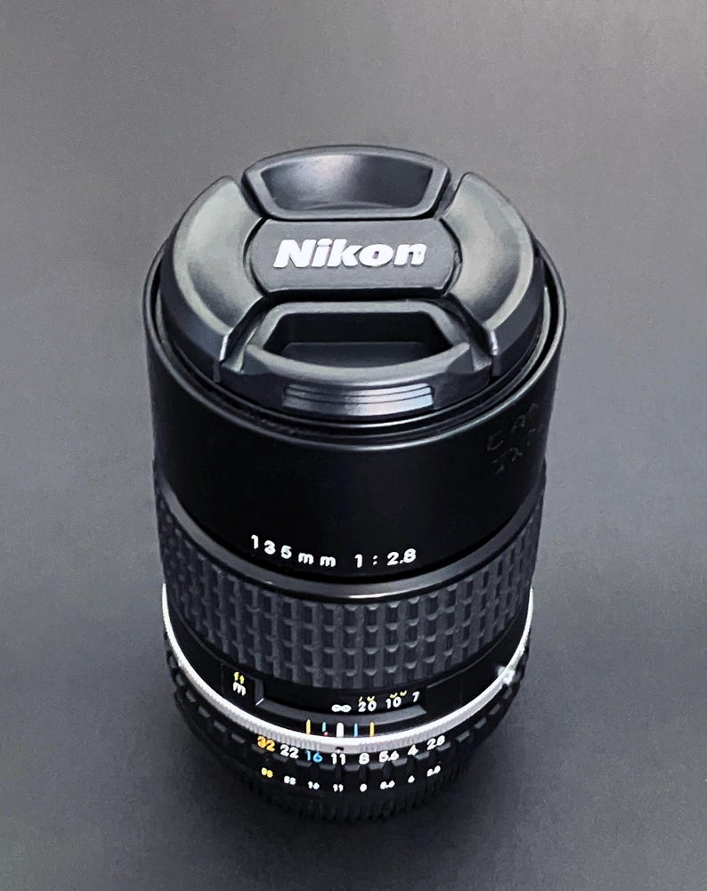 May include: Black Nikon 135mm f/2.8 camera lens with a lens cap. The lens has a ribbed focus ring and aperture settings. The lens cap has the Nikon logo in white.