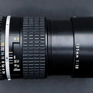 May include: Black camera lens with a 135mm focal length and f/2.8 aperture. The lens has a textured focus ring and a series of aperture settings. The lens is designed for photography and videography.