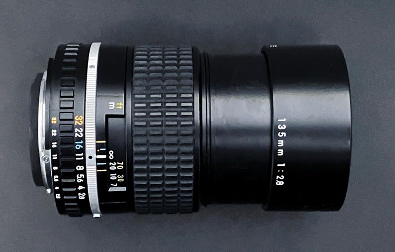 May include: Black camera lens with a 135mm focal length and f/2.8 aperture. The lens has a textured focus ring and a series of aperture settings. The lens is designed for photography and videography.