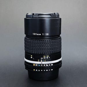 May include: A black camera lens with the text "135mm 1:2.8" printed on the side. The lens has a ribbed grip and a white ring with numbers and markings. The lens is designed for photography.