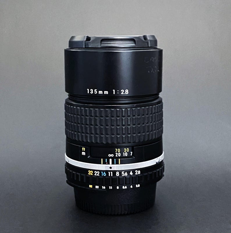 May include: A black camera lens with the text "135mm 1:2.8" printed on the side. The lens has a ribbed grip and a white ring with numbers and markings. The lens is designed for photography.