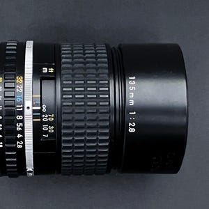 May include: A black 135mm f/2.8 camera lens with a ribbed focus ring and aperture settings. The lens has white and yellow markings, including distance scales in feet and meters. The lens is set against a dark background.