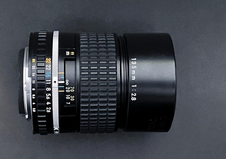 May include: A black 135mm f/2.8 camera lens with a ribbed focus ring and aperture settings. The lens has white and yellow markings, including distance scales in feet and meters. The lens is set against a dark background.