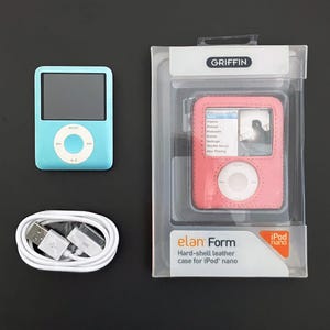 Puede incluir: Un iPod Nano azul claro, una funda de cuero rosa y un cable USB blanco se muestran sobre una superficie oscura. La funda rosa está en un embalaje de plástico transparente con el texto "elan Form" e "iPod nano".