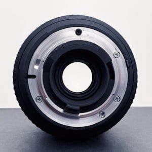 May include: A black camera lens with a silver metal mount. The lens has a large central opening and several small screws around the mount. The lens is designed for use with a camera.