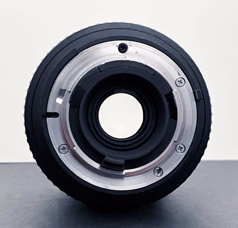 May include: A black camera lens with a silver metal mount. The lens has a large central opening and several small screws around the mount. The lens is designed for use with a camera.