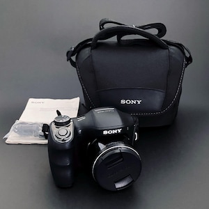 May include: Black Sony Cyber-shot digital camera with a lens cap, a black carrying case, and a manual. The camera has a silver control dial and the Sony logo. The carrying case has a handle and the Sony logo. The camera and case are on a dark surface.