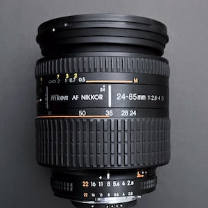 May include: A black Nikon AF NIKKOR 24-85mm 1:2.8-4 D camera lens. The lens has various markings and numbers, including focal lengths from 24mm to 85mm. It features a ribbed design and a focus distance scale.
