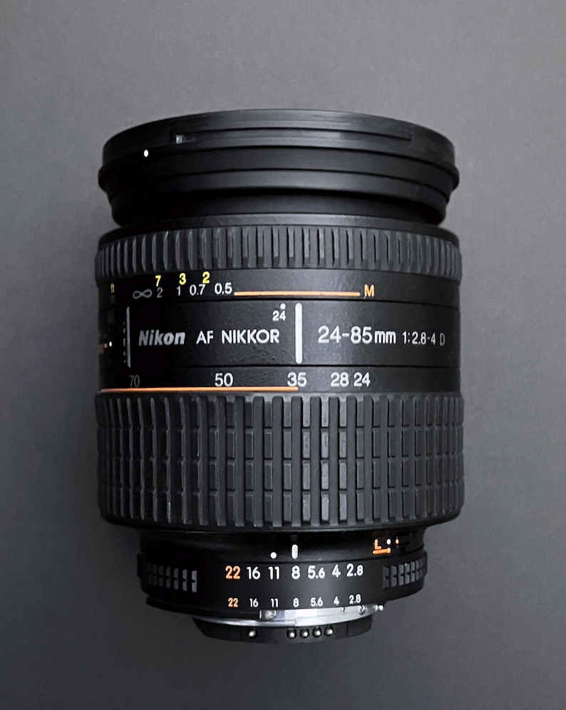 May include: A black Nikon AF NIKKOR 24-85mm 1:2.8-4 D camera lens. The lens has various markings and numbers, including focal lengths from 24mm to 85mm. It features a ribbed design and a focus distance scale.