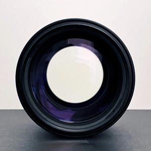 May include: A black camera lens with a large, bright white circular opening in the center. The lens has a dark, reflective interior and a black exterior casing. The image is a close-up, focusing on the lens's front element.