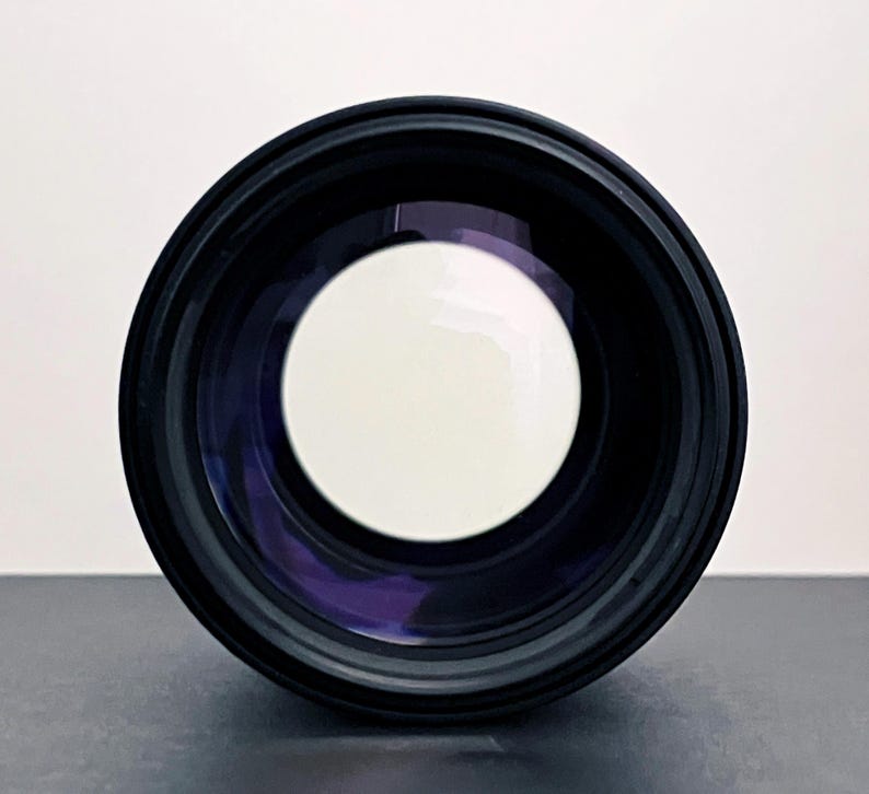 May include: A black camera lens with a large, bright white circular opening in the center. The lens has a dark, reflective interior and a black exterior casing. The image is a close-up, focusing on the lens's front element.