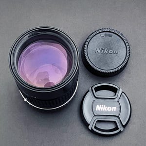 May include: A black Nikon camera lens with a purple lens and two black lens caps. One cap is round with the word "Nikon" and the other is a snap-on cap with the Nikon logo.
