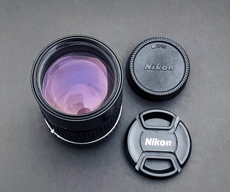 May include: A black Nikon camera lens with a purple lens and two black lens caps. One cap is round with the word "Nikon" and the other is a snap-on cap with the Nikon logo.
