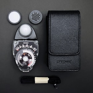 May include: A Sekonic light meter kit, featuring a black and clear meter with a circular dial, two attachments, a black leather case, and a black strap. The meter has the brand name "Sekonic" printed on it.