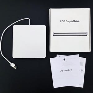 May include: A white USB SuperDrive with a matching white cable and a USB connector. The SuperDrive is next to its white box, which has the product name printed on it. Also included are two white instruction sheets.