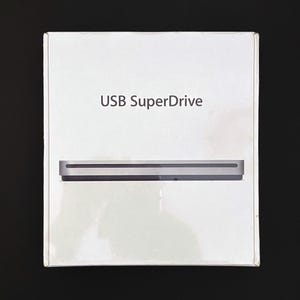 Könnte beinhalten: Eine weiße, rechteckige Produktverpackung mit dem dunkelgrauen Aufdruck USB SuperDrive. Unter dem Text zeigt eine fotografische Abbildung ein flaches, silbernes, metallisches optisches Laufwerk mit einem horizontalen Einzugsschlitz.