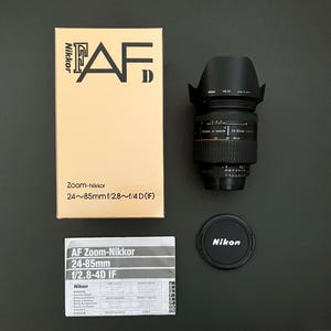 May include: A Nikon AF Zoom-Nikkor 24-85mm f/2.8-4D IF lens kit. The kit includes the lens, lens hood, lens cap, box, and instruction manual. The box is tan with black text. The lens is black with silver markings.