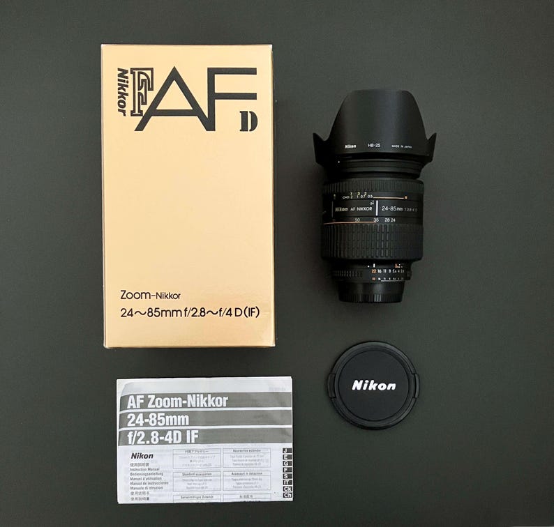May include: A Nikon AF Zoom-Nikkor 24-85mm f/2.8-4D IF lens kit. The kit includes the lens, lens hood, lens cap, box, and instruction manual. The box is tan with black text. The lens is black with silver markings.
