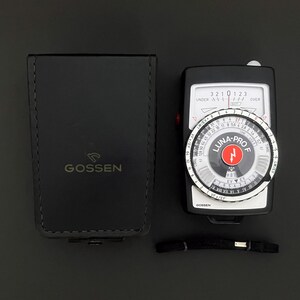 May include: A black Gossen Luna-Pro F light meter with a circular dial, a white display scale, and a red center logo, accompanied by a matching black leather carrying case with the Gossen brand name embossed on the front and a thin black wrist strap.