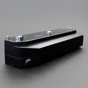 Puede incluir: Accesorio de cámara negro, probablemente una empuñadura o base de batería, con detalles metálicos plateados. El dispositivo rectangular tiene una base curvada y varios tornillos y pasadores plateados en la parte superior. El accesorio está diseñado para acoplarse a una cámara.