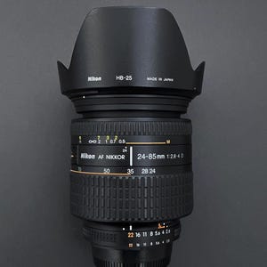 May include: A black Nikon AF Nikkor 24-85mm 1:2.8-4 D camera lens with a lens hood. The lens has markings for focal lengths and aperture settings. The lens hood is labeled "HB-25" and "Made in Japan."