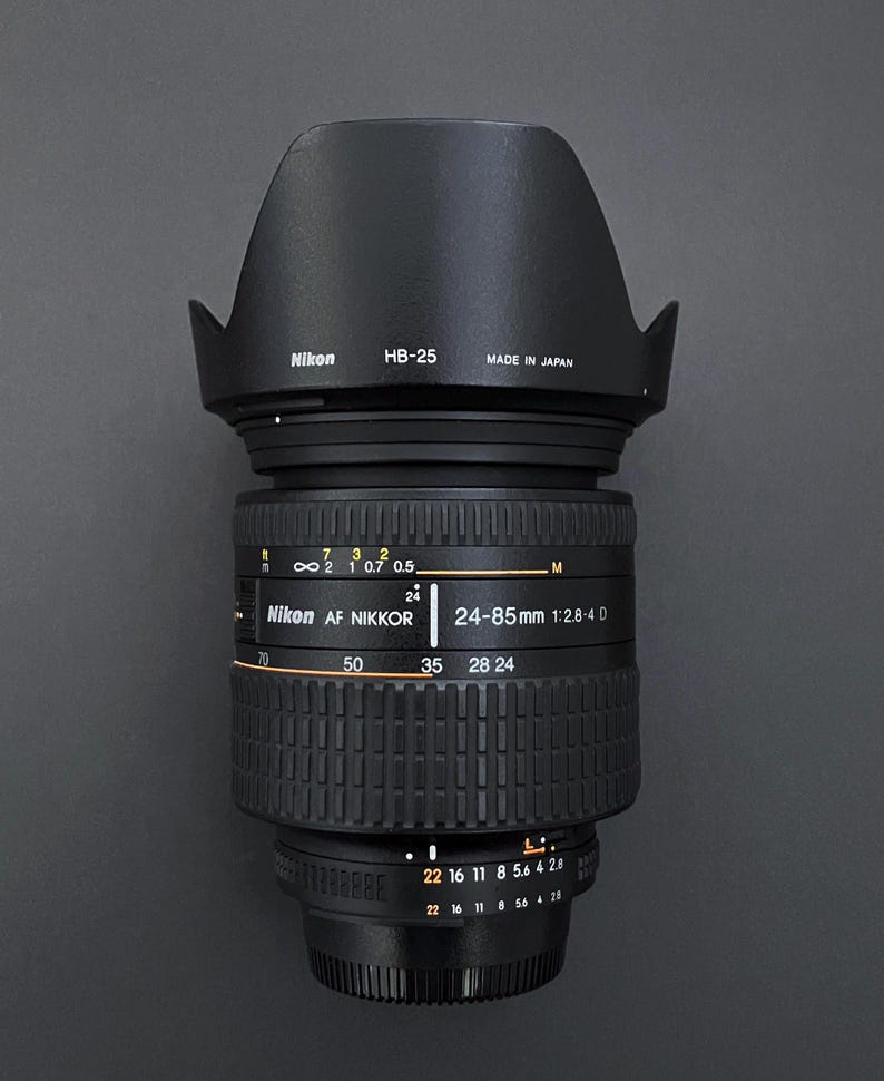 May include: A black Nikon AF Nikkor 24-85mm 1:2.8-4 D camera lens with a lens hood. The lens has markings for focal lengths and aperture settings. The lens hood is labeled "HB-25" and "Made in Japan."
