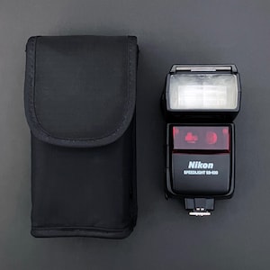 May include: A black Nikon Speedlight SB-600 flash unit with a protective case. The flash has a rectangular head with a white light panel and a red lens. The case is a simple, black pouch.