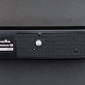 Puede incluir: Accesorio de cámara Minolta Auto Winder G negro con el texto "Minolta Auto Winder G Japan" impreso en la parte delantera.