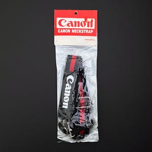 Puede incluir: Una correa para el cuello Canon en un embalaje de plástico transparente. La correa es negra con una raya roja en el centro y el logotipo de Canon en blanco. El embalaje es rojo y blanco con el logotipo de Canon.