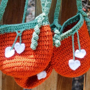 Op de afbeelding: Twee gehaakte tassen met oranje en groene stof. De tassen hebben witte hartvormige versieringen en hangen aan een houten oppervlak.