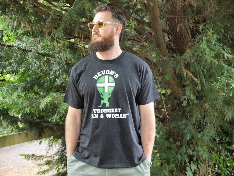May include: A black t-shirt with a white graphic of a person lifting a globe with a green and white cross on it. The text "Devon's Strongest Man & Woman" is printed below the graphic.