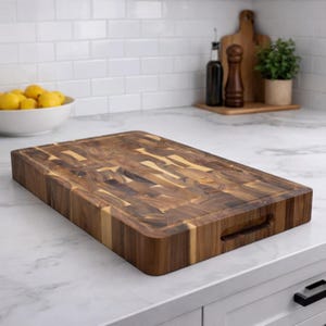 May include: A rectangular wooden cutting board with a dark brown and light brown end-grain pattern. The board has a juice groove around the edge and a handle cutout. It is displayed on a white countertop in a kitchen setting.