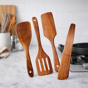 May include: Four wooden kitchen utensils: a solid spatula, a slotted spatula, and two different-shaped spoons. The utensils are made of a light brown wood with visible grain. Each has a hole in the handle for hanging. The background includes a white tile wall and a stovetop.