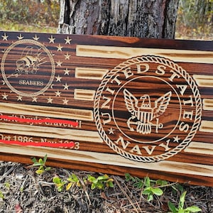 May include: Wooden American flag with engraved details. Features the Seabees logo, the United States Navy seal, and text. The flag is made of dark and light wood, with stars and stripes.