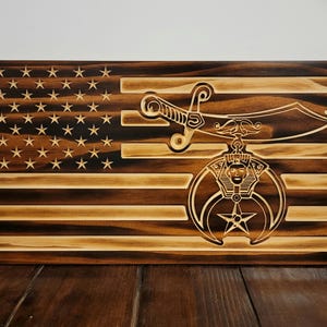 May include: A wooden American flag with a distressed finish. The flag has a 32nd degree Scottish Rite Masonic symbol in the lower right corner. The symbol is a crescent moon with a five-pointed star in the center.