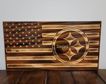 May include: Wooden American flag with the Pittsburgh Steelers logo. The flag features the stars and stripes design, with the Steelers logo in the center. The word "Steelers" is written below the logo.