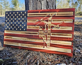 May include: A wooden American flag with a silhouette of a lineman working on a power line. The flag is painted in red, white, and blue with a distressed look.