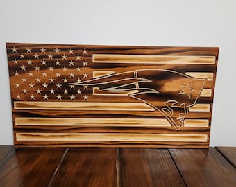 May include: Wooden American flag with the New England Patriots logo. The flag features the stars and stripes design, with the team logo carved into the wood. The wood has a natural, warm tone.