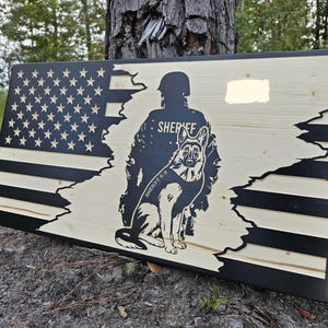 May include: A rectangular wooden sign featuring a black and natural wood American flag design. The center displays a black silhouette of a sheriff in uniform standing behind a German Shepherd dog wearing a vest labeled SHERIFF K-9.